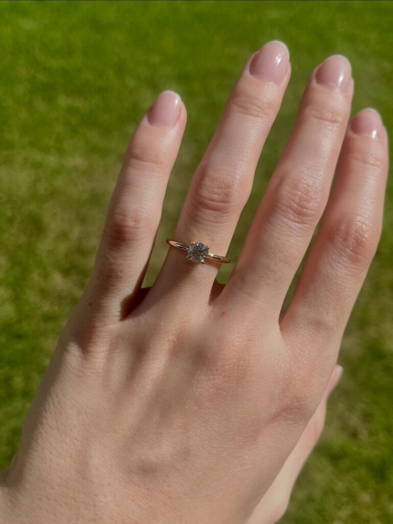 Gold engagement ring with a round diamond on a woman's finger over green grass