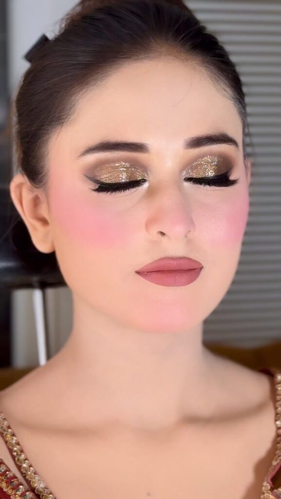 Woman with bridal makeup showing pink blush, gold glitter eyeshadow, and mauve lipstick.
