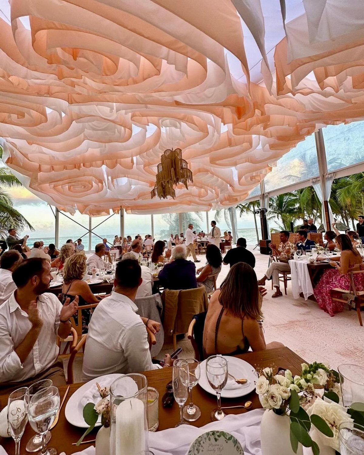 Wedding guests in light attire dining under a flowing peach fabric canopy on a beach.