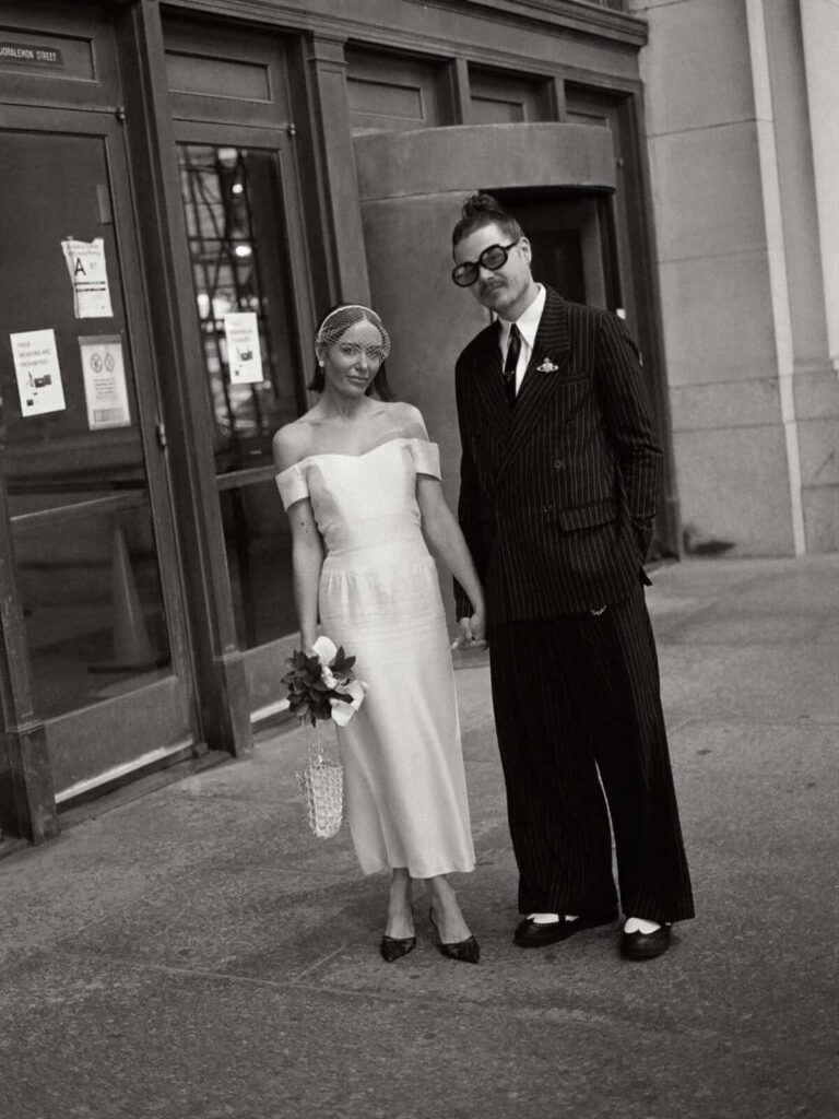 Couple in wedding attire standing outside a city building in black and white.