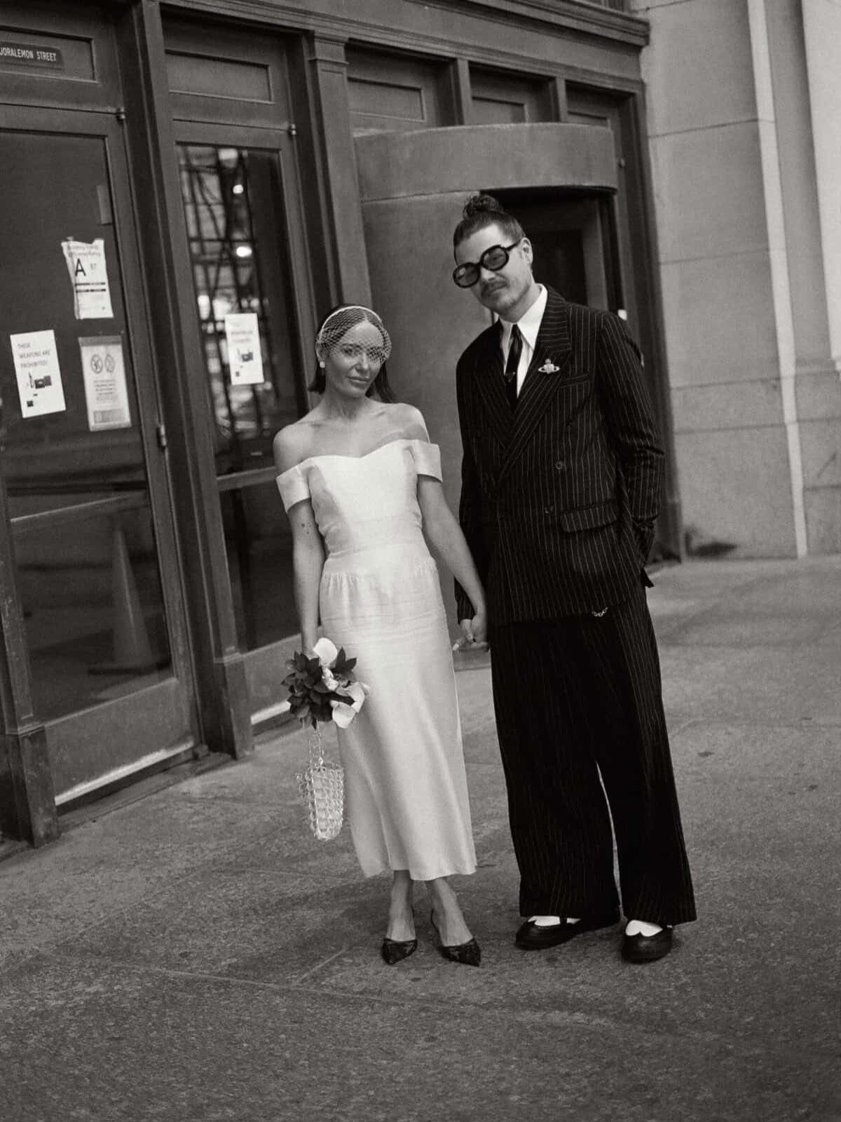 Couple in wedding attire standing outside a city building in black and white.