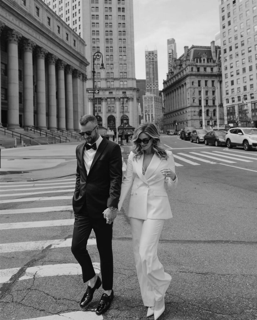 Couple walking in city; woman wearing a white bridal suit and man in tuxedo.
