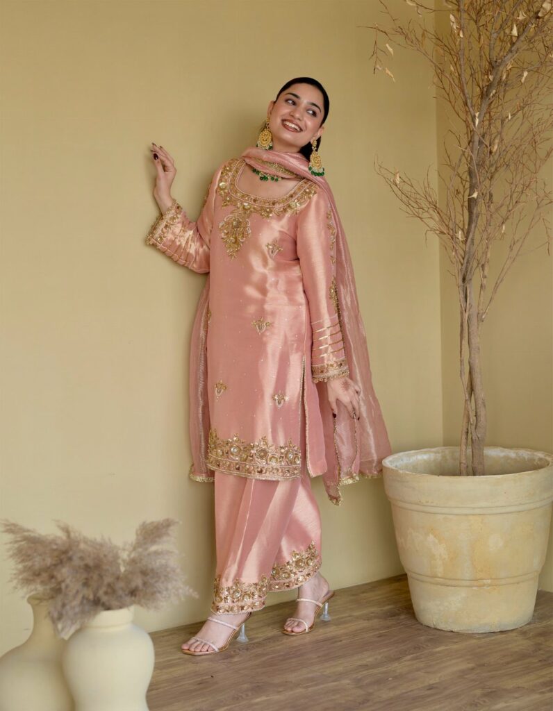 Woman wearing a peach silk salwar kameez with gold embroidery and green earrings.