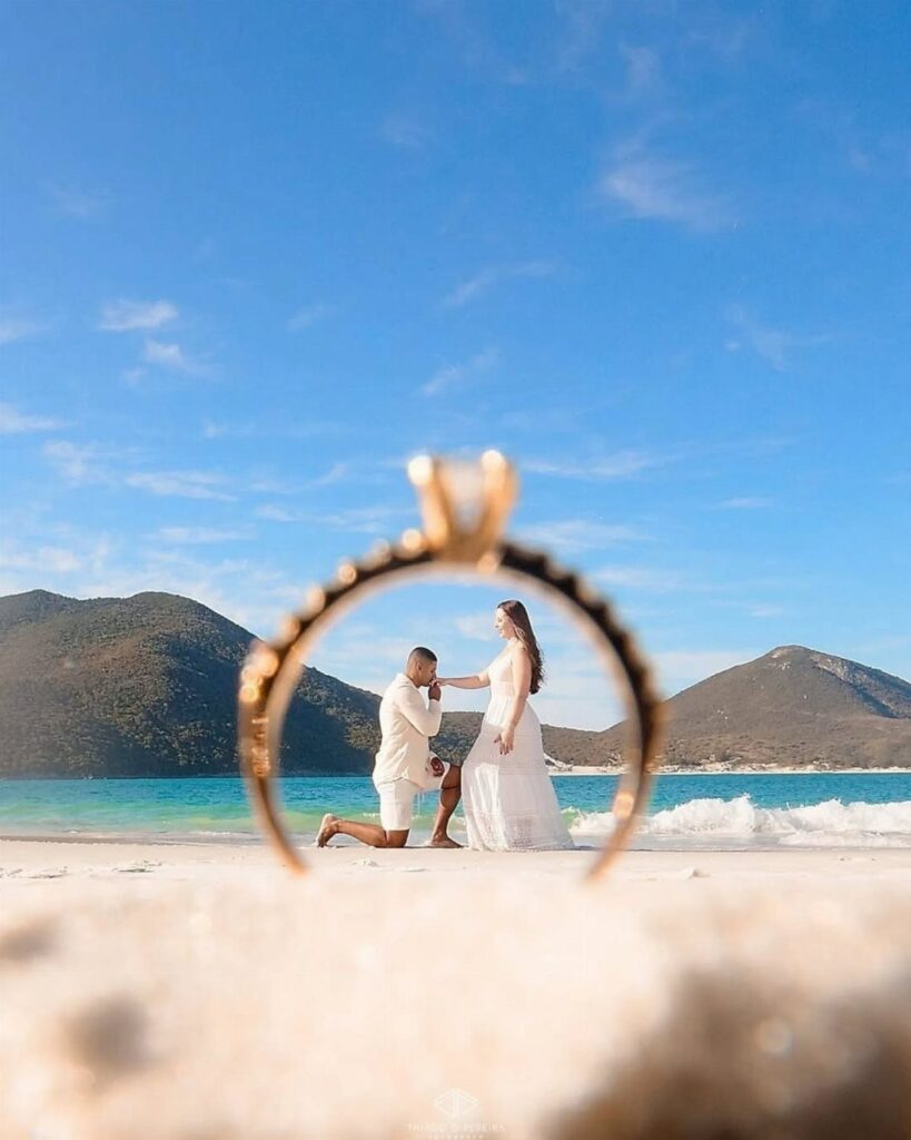 Gold diamond ring framing a man proposing to a woman on a beach