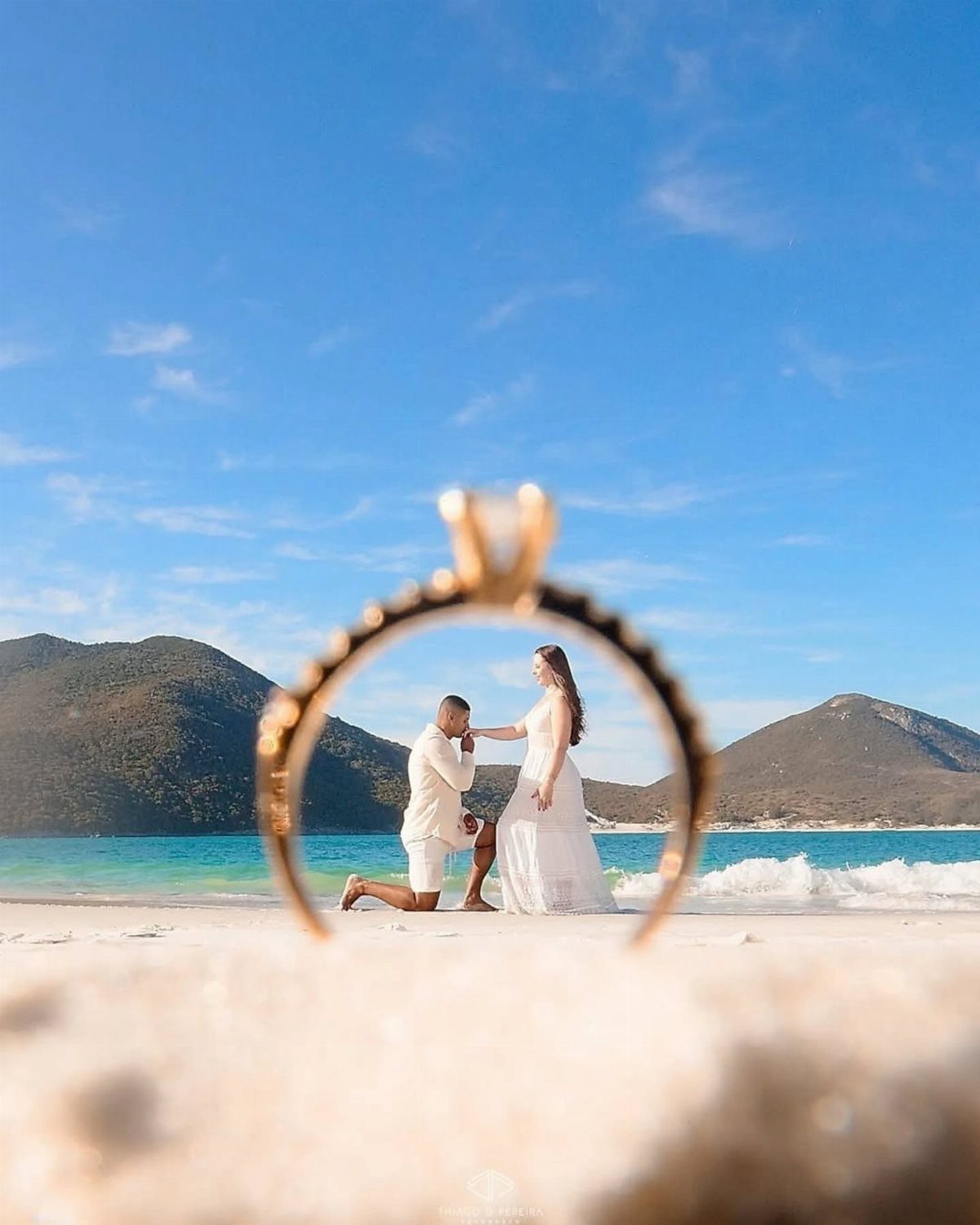 Gold diamond ring framing a man proposing to a woman on a beach