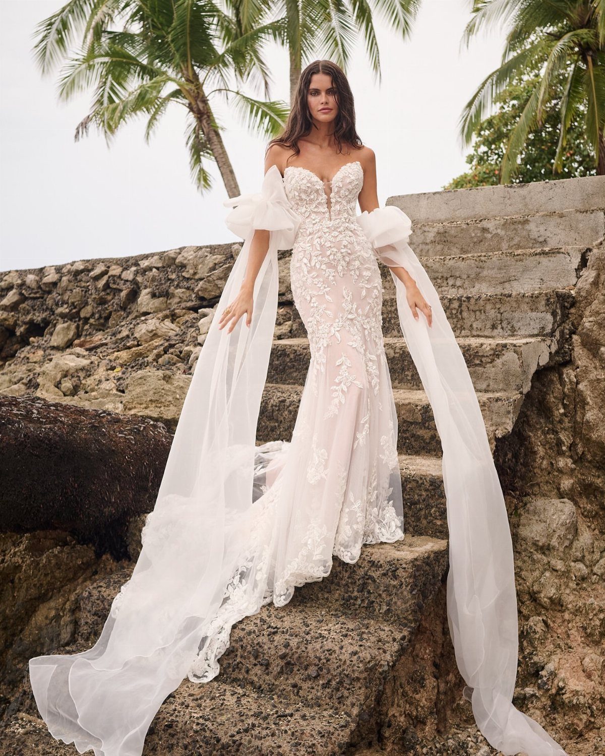 Woman in a white lace wedding dress standing on stone steps with palm trees.