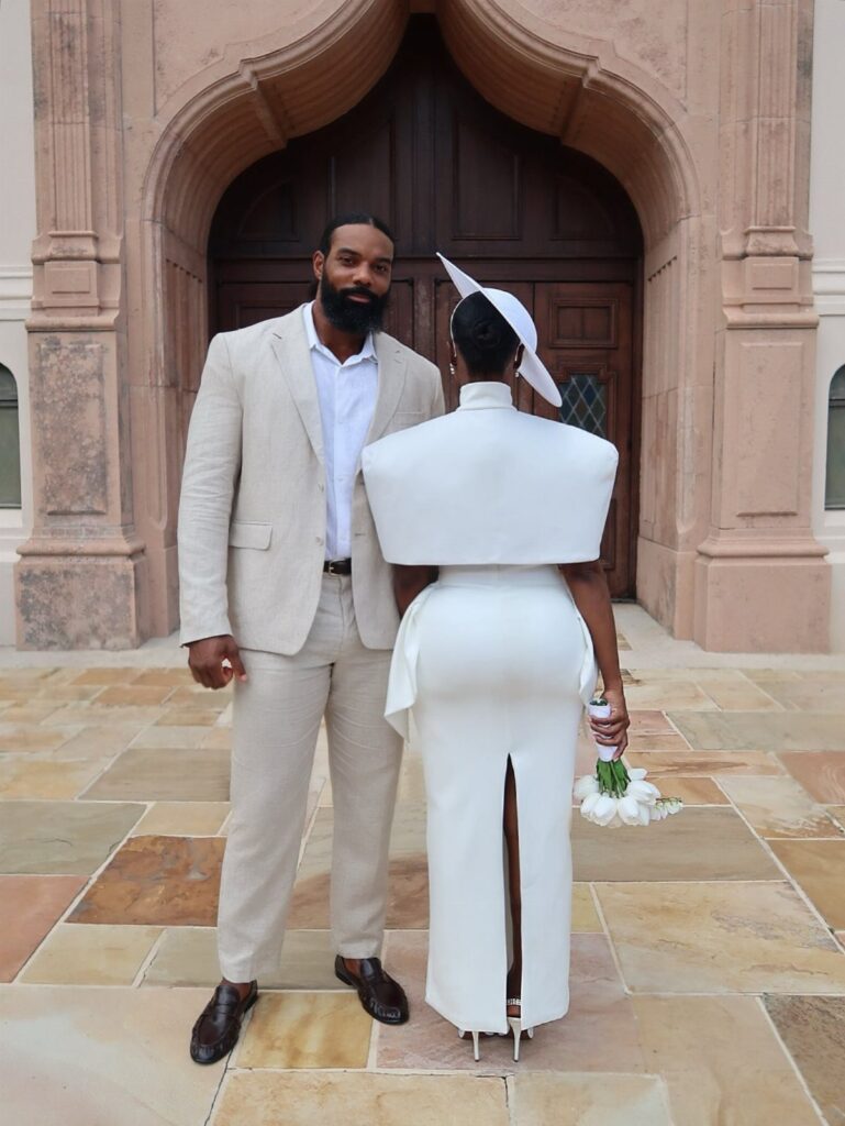Couple in wedding attire standing before a stone archway, woman wearing long white dress.