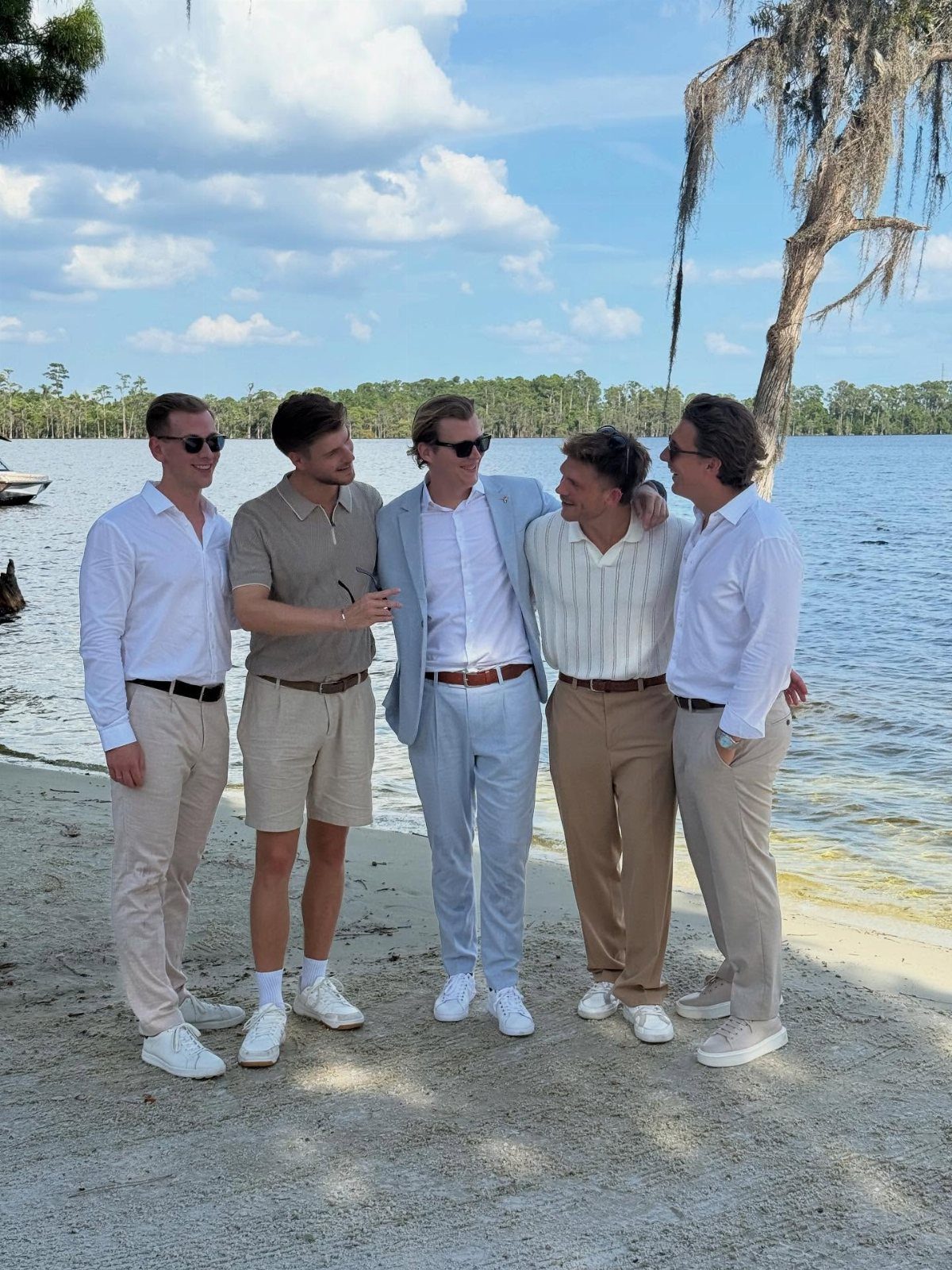 Five men in light-colored summer formal wear standing on a sandy beach lakeside.