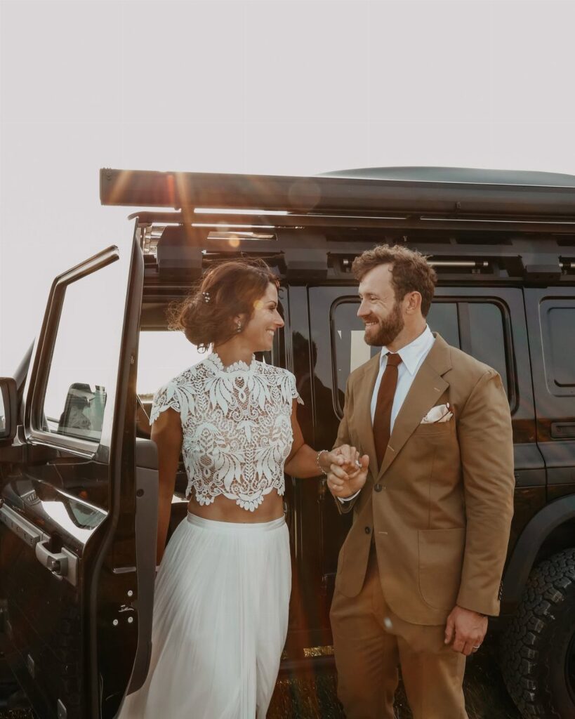 Couple in non-traditional wedding attire posing beside a black off-road vehicle.