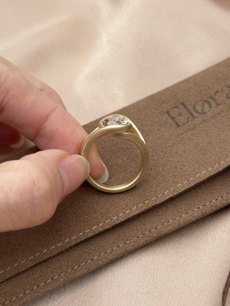 Gold engagement ring with a diamond held over a brown suede pouch.