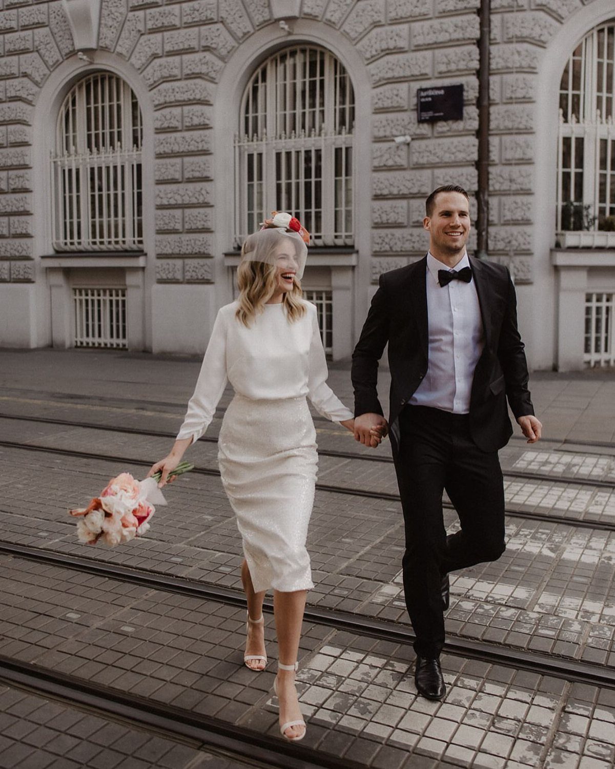 Bride in a simple white midi wedding dress and groom in a tuxedo walking.