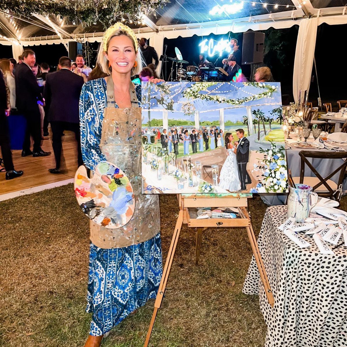 Artist in a paint-splattered apron posing with a wedding painting on an easel.
